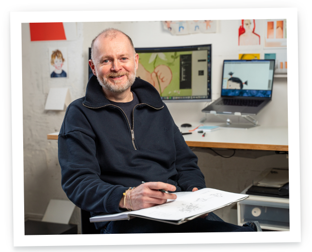An illustrator sitting in his workspace, holding a sketchbook and a pen while smiling at the camera.